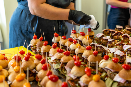 Female Chef Putting Ingredients of Burgers on a Sliced Bread Spread on a Table in Black Gloves - Concept of the Hard Working Person and the Hygiene in the Kitchen, Partly Blurred Backgroundの写真素材