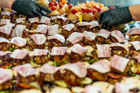 Female and Male Chef Putting Ingredients of Burgers on a Sliced Bread Spread on a Table in Black Gloves - Concept of the Hard Working Person and the Hygiene in the Kitchen, Partly Blurred Backgroundの写真素材