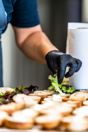 Female Chef Putting Ingredients of Burgers on a Sliced Bread Spread on a Table in Black Gloves - Concept of the Hard Working Person and the Hygiene in the Kitchen, Partly Blurred Backgroundの写真素材