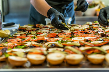 Female Chef Putting Ingredients of Burgers on a Sliced Bread Spread on a Table in Black Gloves - Concept of the Hard Working Person and the Hygiene in the Kitchen, Partly Blurred Backgroundの写真素材