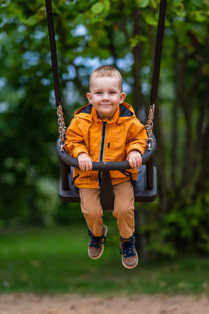 Young boy wearing orange raincoat smiles in swings in the parkの写真素材