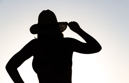 Backlit silhouette of a woman holding a hat with one hand.の写真素材