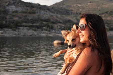 Young woman with her Pomeranian dog in her arms with a lake in the background.の写真素材