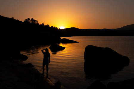 Woman on the shore of a lake at sunset. Overcoming life's difficulties. Concept of reflection and thought. Selective focus. Copy space. Self-confidence.の写真素材