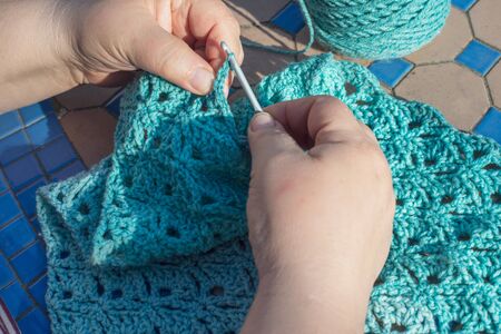 Woman making a blue shawl or scarf with crochet hook in garden at home.