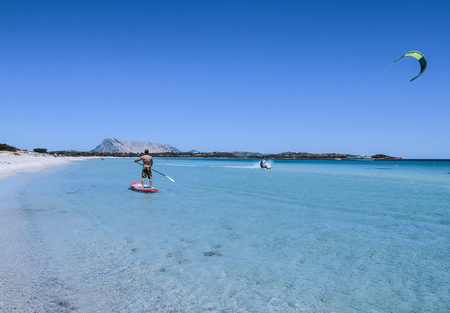 San Teodoro - La Cinta Beach - 2017 July - A young athlete paddle on stand up paddle on the crystalline sea of Sardinia, while a kitesurfer slides on the water beside himのeditorial素材