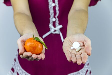 Vitamins from fruits or medicines? A young woman in burgundy pajamas shows a mandarin in her right hand and an aspirin in her leftの写真素材