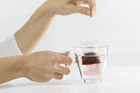 Close-up of a woman hand dipping a sachet of Hibiscus Tea in a glass cup full of water with...