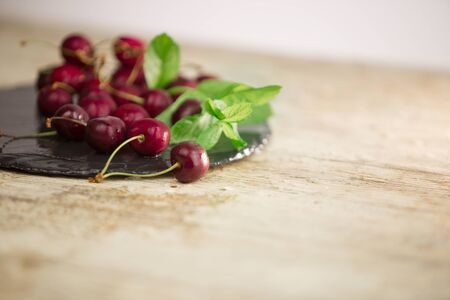 Fresh cherries on a black plate of wet slate with sprig of fresh green mint on light wooden table in selective focus for copy spaceの写真素材