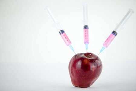 Concept: human GMO manipulation of nature and relative poisoned fruits. Close-up of an apple contaminated with three syringes threaded in and white backgroundの写真素材