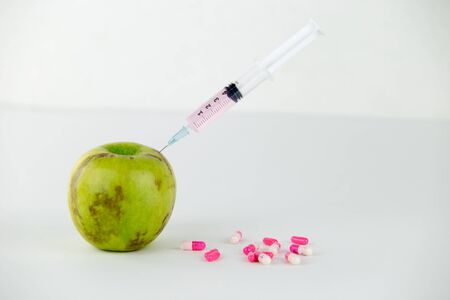 Concept: human GMO manipulation of nature and relative poisoned fruits. Close-up of an apple contaminated with a syringe threaded in and medicines on a white backgroundの写真素材