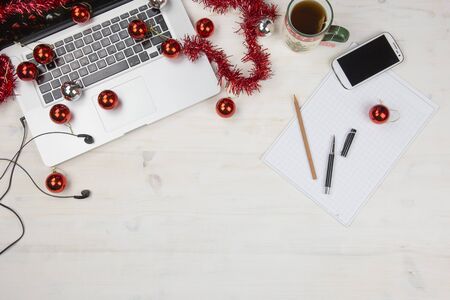 Job at Christmas holidays: flat lay of a light wooden table with an open aluminum laptop, red decoration, red baubles, Christmas cup of tea, smartphone, block notes, pencil and pen on block noteの写真素材