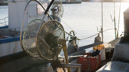 A moored fishing boat is waiting to set sail water while the sun reflects on the sea early in the morning with the winch of the fishing net in the foregroundの写真素材