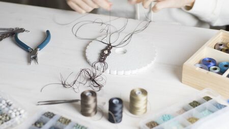 Lifestyle concept, work from home to reinvent your life: close-up of woman hands making macrame knotted jewelry with stone beads and tools on light wooden tableの写真素材