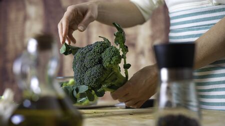 Moments of daily life in Mediterranean cuisine: low angle view of a young female cook cleans broccoli on a wooden chopping board with extra virgin olive oil, garlic and the knife with bokeh effect.の写真素材