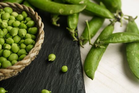 Macro close up low angle view of fresh shelled peas in a wicker basket on a black slate plate and some pods on a light wooden background. Black, white and green contrast from healthy vegan cuisineの写真素材