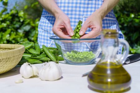 Close up of the hands of a woman shelling peas from the pod inside a glass bowl outdoors with sunlight in the greeneryの写真素材