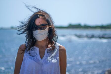 Coronavirus seaside holidays: half-length shot of a woman at the beach with the mask for Covid-19 pandemic with cloudy skyの写真素材