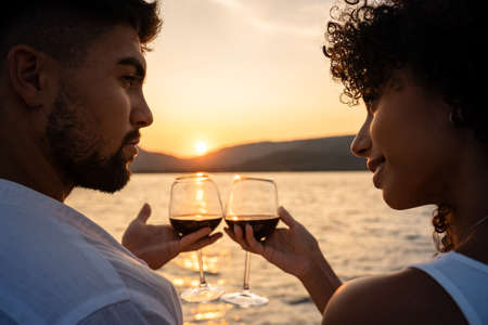 Romantic close-up of a mixed race couple toast with glasses of red wine crossed by the light of the setting sun on a lakeの写真素材