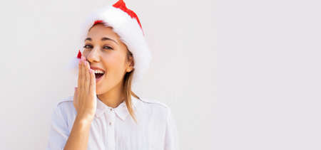 Young beautiful caucasian blonde woman with a Santa Claus hat surprising with his hand in front of...