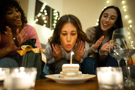 Close up of cute young woman blows on the cake candle next to her present celebrating her birthday...
