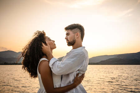 Attractive mixed race lovers couple in romantic pose in sunset backlight - Strong serious bearded young handsome man holding face of her black Hispanic girlfriend looking in her eyes with passionの写真素材