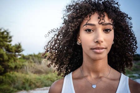 Femininity and beauty in nature: close up portrait of beautiful black Hispanic young woman with curly dark long hair looking confident at the camera - Make-up female artist with perfect mouth and eyesの写真素材