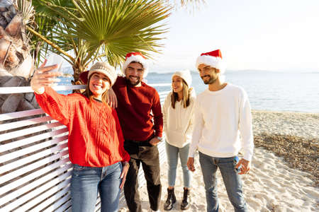 Group of young caucasian friends with Santa Claus hat having fun taking a selfie at the beach in winter for the Christmas holidays - Two couple of people using technology to sharing their journeyの写真素材