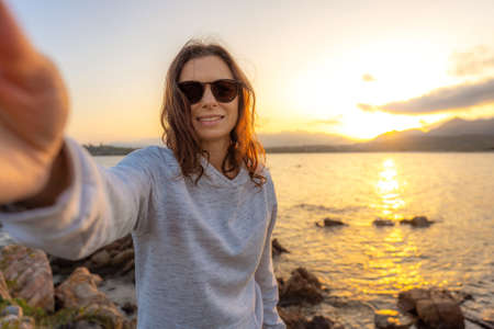 Young beautiful caucasian woman make a self portrait holding the camera at sunset on sea rocks -...