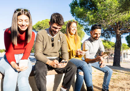 Two beautiful young couple sitting on a wall having fun using smartphone - Friends group smiling...
