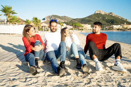 Two young couples by the sea at sunset in a seaside resort in winter having fun talking and laughing each other - Group of friends sitting on the beach in winter sea vacation - White and red clothingの写真素材