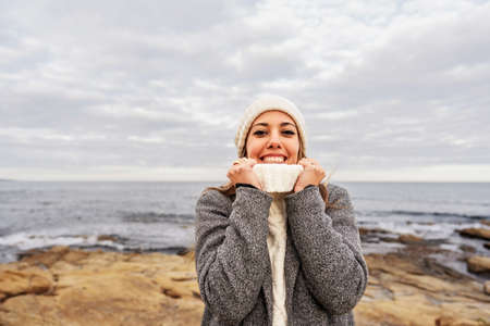 Beautiful young Caucasian woman tightens the neck of her white sweater with her hands for the cold...