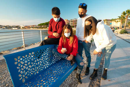 Two Caucasian heterosexual couple in red and white sweaters looking at the smartphone in vacation winter resort - Group of friends wearing protection mask using mobile technology to planning journeyの写真素材