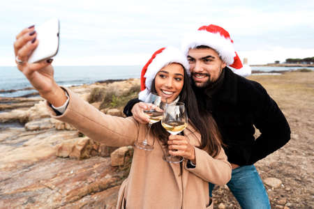 Young beautiful multiracial couple making a self portrait outdoor in sea resort wearing Santa Claus hat holding white wine glasses celebrating Christmas holidays and new year event in winter vacationの写真素材