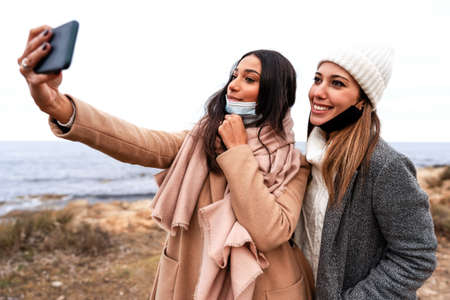 Beautiful stylish Hispanic woman making selfie outdoor in sea winter vacation with her Caucasian...