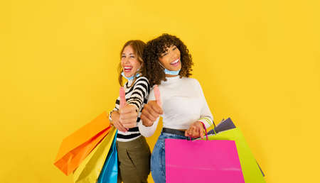 Two confident mixed-race young women holding colored shopping bags and smiling doing thumbs up looking at the camera on big yellow background copy space, wearing Coronavirus protection maskの写真素材