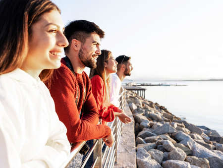 Young beautiful happy people leaning on a metal railing at the sunset on the water in sea winter vacation resort - Confident friends group looking at the horizon - Focus on closer bearded manの写真素材