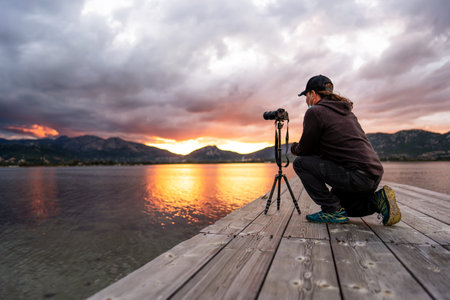 A tourist keen on photography bends down to adjust his camera to get a good picture of the spectacular cloudy sunset with the sun between the mountains and the orange reflection of light in the waterの写真素材