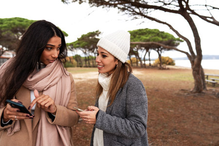 Beautiful latina young woman pointing her cell to showing photo to her Caucasian girlfriend outdoor - Lesbian cute mixed race couple having fun outside in sea resort using social network on smartphoneの写真素材