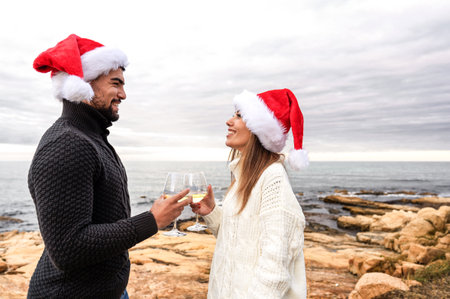Funny romance scene of a beautiful happy young couple celebrating Christmas standing outdoor face to face wearing Santa Claus hat toasting with champagne glasses in natural sea resort winter vacationの写真素材