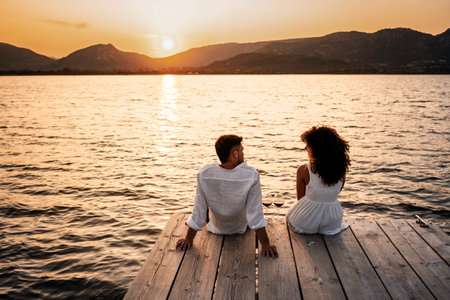 Romantic scene of couple in love at sunset sitting on wooden jetty with wine glasses - Hispanic curly brunette sitting on a pier of lake with orange water reflection looking to her handsome boyfriendの写真素材
