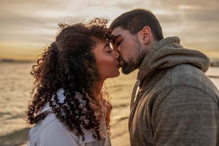 Head and shoulders portrait of young beautiful couple in love kissing at sunset in winter seaside resort with cloudy sky. Two millennials in vacation travel manifesting their heart sentiments outdoorの写真素材