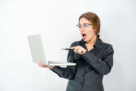 Surprised isolated beautiful young woman posing in study shot holding laptop in hand pointing with pen the screen wearing office business clothes. Caucasian female employee in wow expressions watch pcの写真素材