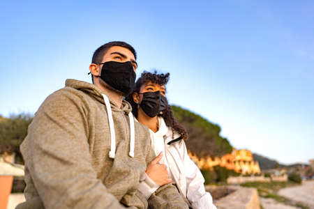 MIxed-race afro-american girl embracing her Caucasian boyfriend sitting outdoor in sea resort looking the sunset wearing black protective mask against Coronavirus pandemic. New normal vacation travelの写真素材