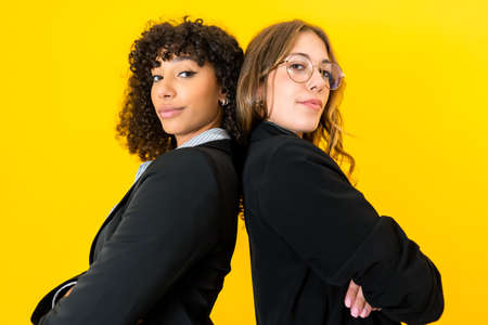 Confident multiracial business women back to back with crossed arms looking at camera isolated on yellow background for copy space. Female success on work and better life conceptの写真素材