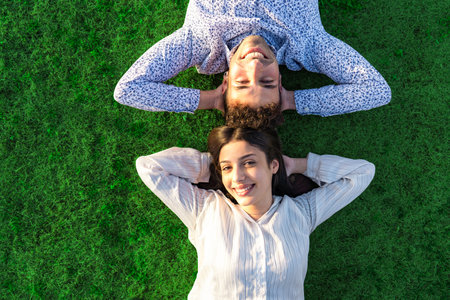 Happy young couple view from top with crossed arms under head smiling looking at camera lying on a green grass meadow with sunset or dawn colored light. Concept of confident people trust in the futureの写真素材