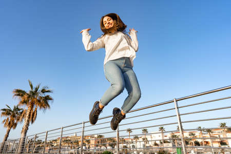 Happy smiling girl jumping outdoor high with arms up looking down at the camera with her hair ruffled by the wind. Concept of happiness and good life emotion for success of positive people in youthの写真素材