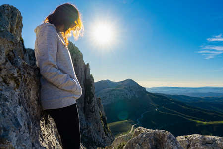 Lonely woman finds herself leaning against the rocks in the nature pensive at top of mountains with a splendid view below and sun through her hair. Concept of positive loneliness to find your own wayの写真素材