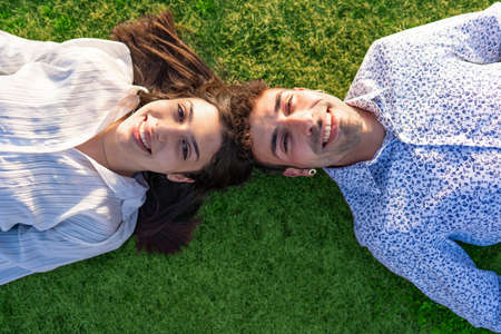 Young couple of beautiful people lying on the ground on a green lawn at sunset or sunrise head to head looking at camera from below laughing happily and confidently in the future. Concept of wellbeingの写真素材