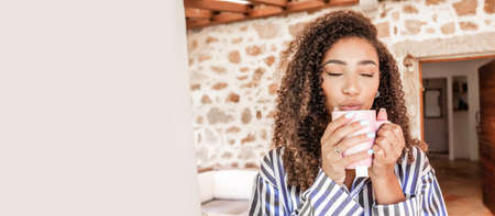 Beautiful black Hispanic girl with dark curly hair in striped silk pajamas enjoying her cup of hot tea holding it in her hands with her eyes closed on the porch of her country house. Left copy spaceの写真素材